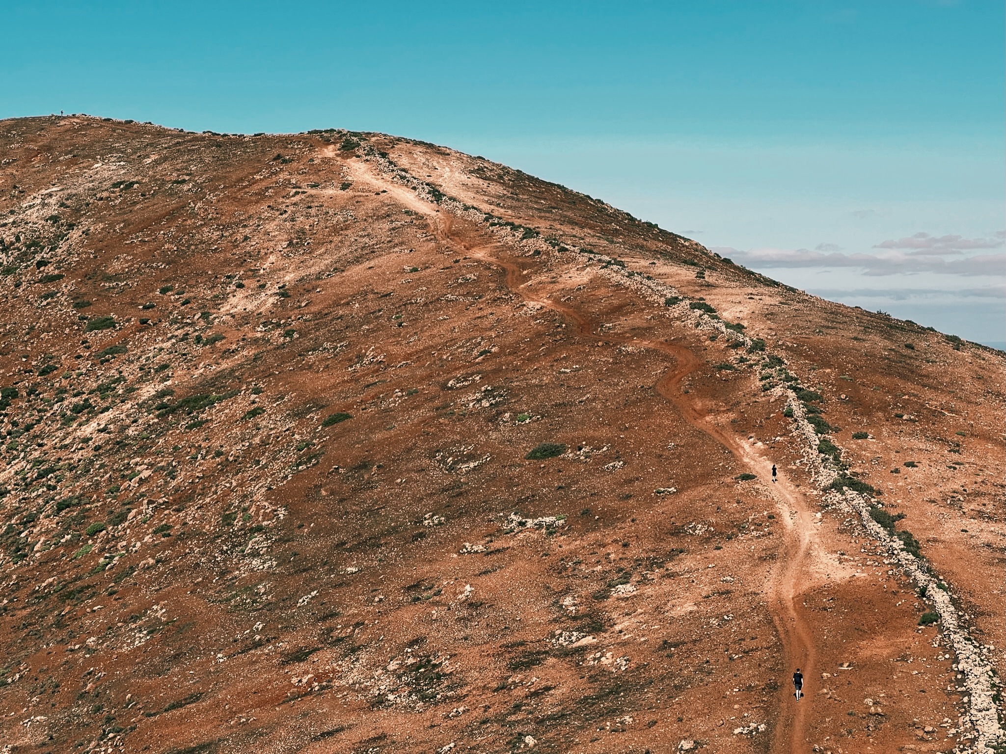 Two people hiking along long red gravel road up a large hill on Fuerteventura, Spain