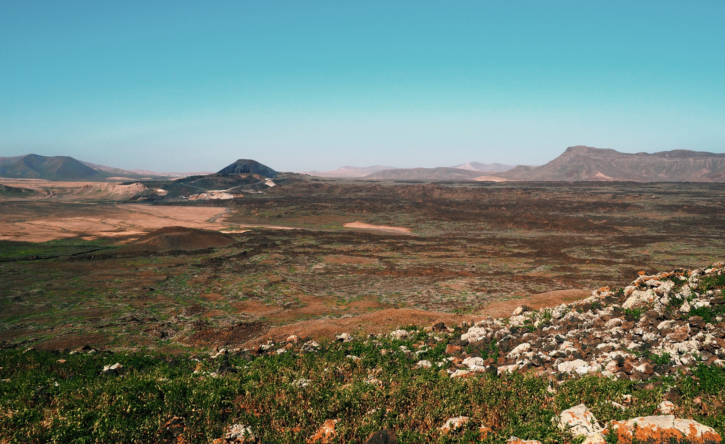 View over broad landscape and volcanic gravel in hues of green, brown, and orange leading to Caldera Los Arrabales, a hiking route on Fuerteventura